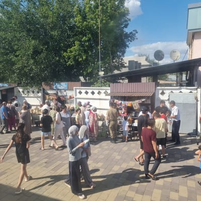 The image depicts a bustling outdoor market scene, likely in Turkistan, Kazakhstan, on a sunny day. Several stalls are set up, displaying various goods including packaged snacks, drinks, and produce such as watermelons and other fruits. A group of people are browsing the stalls and interacting with vendors. The architecture suggests a blend of modern and traditional elements, with corrugated metal roofing and simple stall structures. The ground is paved with interlocking bricks. The sky is bright blue with scattered clouds, and lush green trees are visible in the background, providing shade. The overall atmosphere is one of daily commerce and community activity.