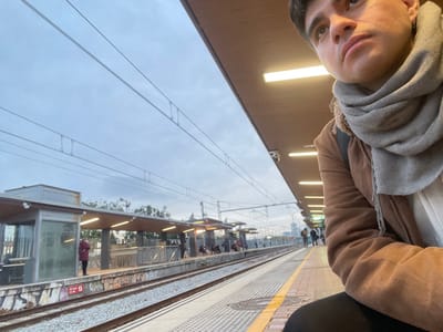 A person wearing a tan jacket and grey scarf stands on a train platform. Other people wait in the distance. A metal railing separates the platform from the train tracks, which are covered in graffiti. Overhead power lines stretch across the grey sky. The platform is tiled and lit by fluorescent lights. The individual in the foreground looks off to the side.