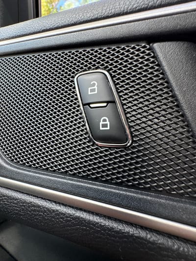 The image shows a close-up view of a car's interior door panel, specifically the lock and unlock buttons on the speaker grill. The buttons are black with white icons. The top button features an unlocked padlock symbol, and the bottom button shows a locked padlock symbol. The speaker grill has a metallic mesh texture. The surrounding door panel is black with a textured finish and a silver trim strip. The lighting suggests this is an indoor environment, possibly a garage or a car parked in daylight. There is no indication of the location being in Мазалаўскі сельскі Савет, Belarus, or any other specific city.