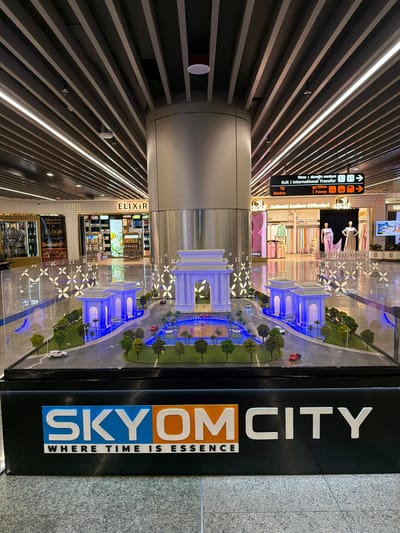 In the city of Vikram Nagar, India, this image captures a detailed architectural model displayed within what appears to be a modern airport or large public transport hub. The model, centrally positioned, showcases a miniature cityscape with an iconic archway, flanked by smaller structures and illuminated with vibrant blue lights. The surrounding area features miniature trees, roads with toy cars, and a central water feature, all enclosed within a clear protective barrier. In the background, shops like "ELIXIR WINE & SPIRITS EMPORIUM" and "ada Authentic Lucknow Chikankari" are visible, indicating a commercial setting within the transportation hub. Overhead, a distinctive ceiling design with parallel beams and integrated lighting runs across the space. A directional sign with English and Hindi text indicating "Exit | International Transfer," "Metro," and "Forex" further confirms the airport or major transit station environment. The overall scene is well-lit, emphasizing the display model and the surrounding amenities.