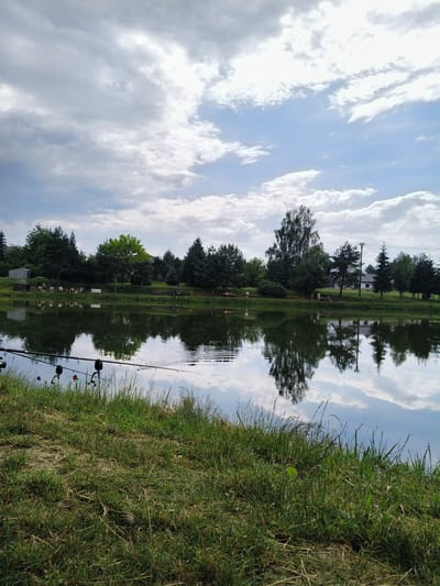 This image depicts a serene outdoor scene at a lake, likely in Ćwiklice, Poland. The primary subjects include a body of water, a grassy bank with several fishing rods set up, and a treeline on the opposite shore. In the background, there are several people gathered, some sitting and others standing, suggesting a leisurely fishing activity or a social gathering by the lake. A few structures, possibly small buildings or sheds, are visible amongst the trees, along with a white bench.

The time of day appears to be late afternoon or early evening, judging by the lighting and the soft reflections on the water. The weather seems partly cloudy, with dramatic formations of white and gray clouds against a blue sky, creating a dynamic contrast. The reflections of the trees, clouds, and people are clearly mirrored on the calm surface of the lake, adding depth and symmetry to the composition.

Notable details include the presence of multiple fishing rods positioned on the bank, indicating that fishing is the main activity. The overall emotion conveyed is one of peace and relaxation, a common feeling associated with nature and outdoor leisure. The lush green grass in the foreground and the dense foliage of the trees on the far bank contribute to the natural and tranquil atmosphere of the location. There is no visible text in the image.