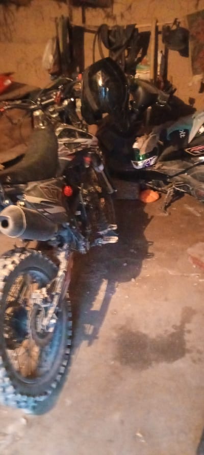 The image depicts a cluttered garage or workshop interior. The primary subject is a dirt bike, positioned centrally and angled towards the viewer. The bike appears to be a dual-sport or enduro model, with knobby tires suggesting off-road capability. It is dark in color, with some metallic accents and branding visible. A black motorcycle helmet with green and purple accents rests on top of some other items behind the bike, possibly indicating it's being stored or worked on.
The background is filled with various objects, including what appear to be other motorcycle parts, tools, and miscellaneous items hanging on the wall or piled on shelves. The walls seem to be made of rough, textured material, possibly brick or concrete. The floor is a concrete surface, with some dirt and debris scattered around. Shadows are cast on the floor, suggesting artificial lighting from above or to the side, typical of an indoor setting. The lighting is dim, contributing to a somewhat murky atmosphere, and the overall impression is one of a functional but untidy workspace. There is no visible indication of the time of day or weather conditions outside, as the scene is entirely indoors. No people are visible in the image. The location context provided is Бетқайнар, Kazakhstan, which suggests this is likely a private garage or workshop in that region. There is no discernible text within the image itself, beyond potential manufacturer logos on the motorcycle or helmet, which are too blurry to read.