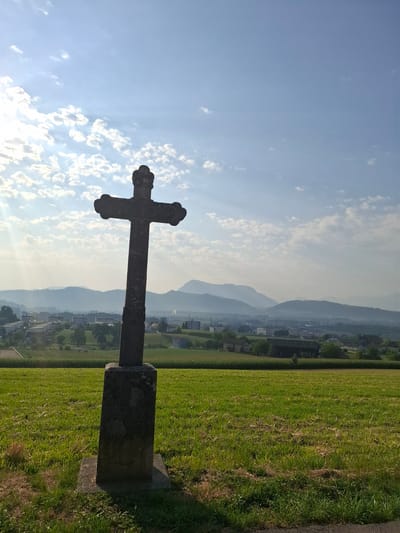 A stone cross stands in the foreground on a grassy hill, silhouetted against a bright sky. The cross is dark and weathered, with a detailed, ornate top. The base of the cross is a rough, rectangular stone structure. The hill slopes down towards a valley where a town is visible, with buildings and houses scattered amongst trees. Beyond the town, rolling hills and mountains recede into the hazy distance. The sky is blue with scattered white clouds, and sunlight streams from the upper left, casting a shadow from the cross onto the grass. The environment is rural and peaceful, with a sense of elevation and a panoramic view. There are no people visible in the image. The specific location is on a hill overlooking the municipality of Eschenbach (LU) in Switzerland.