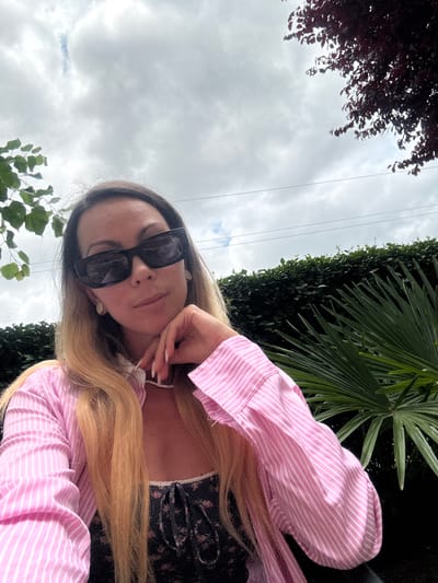 A woman with blonde hair and sunglasses is posing for a selfie outdoors. She is wearing a pink and white striped button-down shirt over a dark floral top. Her left hand is resting on her chin, and she is looking at the camera. In the background, there is a hedge, a palm frond, and a cloudy sky. The image appears to have been taken in Kosharitsa, Bulgaria.