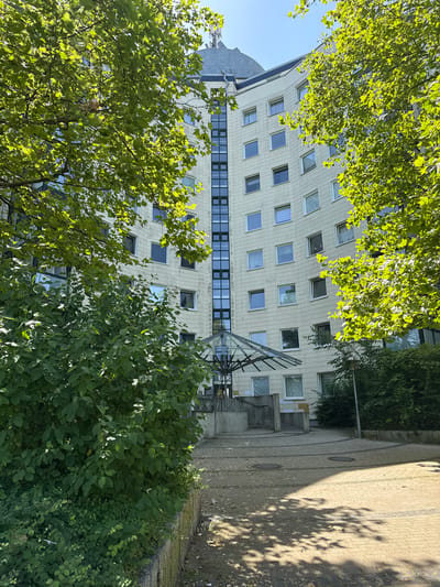 This image shows an exterior view of a curved, modern apartment building in Wolfsburg, Germany, likely located in the Nordstadt district, given the architectural style and surrounding greenery. The building's facade is composed of light-colored panels with numerous rectangular windows. Green trees with large leaves frame the building from the left and right, casting dappled shadows on the paved courtyard in the foreground. A covered entrance with a glass roof is visible in the center of the frame, leading into the building. The courtyard is paved with interlocking bricks arranged in concentric circular patterns. The overall impression is one of a residential area with a focus on integrating nature into the urban landscape. There are no people visible in the image.