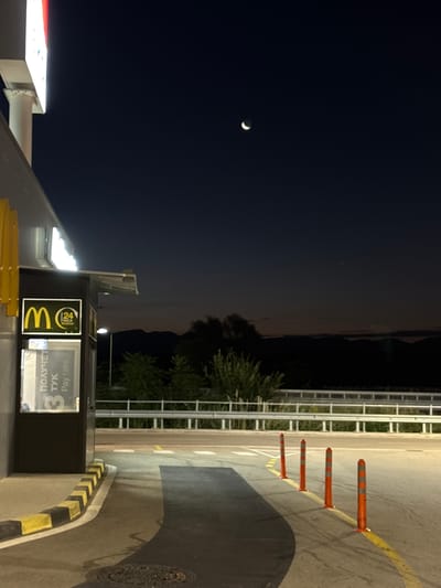 This is a night-time photograph of a McDonald's drive-thru in Chuchuligovo, Bulgaria. The image captures a dimly lit scene with a crescent moon visible in the dark sky. The McDonald's building is on the left, with its illuminated signage and a service window displaying the McDonald's logo and text in what appears to be Bulgarian and English ("Pay here"). Orange traffic cones are lined up on the right side of the paved drive-thru lane. In the background, a metal railing separates the drive-thru from a darker area with trees and silhouetted hills or mountains against the horizon. The overall atmosphere is calm and quiet, characteristic of late-night operations.