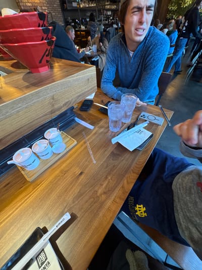 The image depicts an indoor scene at Jinya Ramen Bar, located at 1233 N Eddy St, South Bend, Indiana, United States.
In the foreground, a polished wooden counter serves as a dining surface. On the counter, from left to right, are: a black rectangular plate holding a menu and a white chopstick wrapper with social media handles; a small wooden tray containing four white ceramic condiment containers labeled "GYOZA SAUCE," "CHILI OIL," and "SPICY SAUCE," along with an empty dipping dish; a clear plastic stirrer; a second white chopstick wrapper.
Seated at the counter on the right is a male individual with a distinct facial expression of discomfort or intense reaction, characterized by squinted eyes and a partially open mouth. He wears a blue long-sleeved quarter-zip shirt and a grey baseball cap. In front of him are two clear glasses filled with ice water, a black smartphone displaying "10:04", a white smartphone in a clear case, and a pair of wooden chopsticks.
The lower right foreground shows a partial view of another individual's arm and leg. This person wears grey sweatpants and a navy blue t-shirt with a yellow Notre Dame "ND" logo and "MEN'S CLUB SPORTS" text. A clenched fist is visible.
The background reveals more of the restaurant interior, including a stacked display of three red ramen bowls with black Japanese characters on a wooden shelf. Beyond this, patrons are seated at tables and at a well-stocked bar. The decor features brick walls and dark ceiling elements, contributing to a modern restaurant atmosphere.