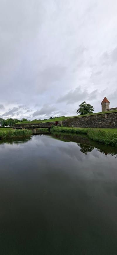 This media file depicts a scene of a fortification with a prominent tower, reflecting in calm water. The fortification is constructed of stone and topped with lush green grass. A conical-roofed tower with a small pointed spire stands to the left, its reddish-brown hue contrasting with the muted colors of the cloudy sky. Trees and grassy slopes line the top of the fortification, creating a naturalistic border.
The water in the foreground acts as a mirror, reflecting the fortification, the sky, and the surrounding greenery. The reflection is slightly distorted, indicating the stillness of the water's surface. A pathway or boardwalk runs along the edge of the fortification next to the water, with some railings visible. A few figures, appearing as small red dots, are present on this path, suggesting human presence and activity within the historical site. The overall mood is serene and somewhat overcast, with the gray clouds suggesting a possibility of rain or a generally cooler day. The time of day is indeterminate, but the lighting suggests it is daytime. The location is identified as Kuressaare, Estonia, known for its well-preserved medieval castle, which this fortification and tower likely belong to. There is no visible text in the image.