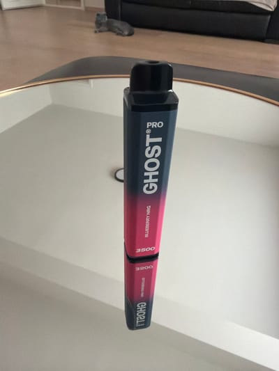 The image shows a Ghost Pro blueberry NRG vape pen positioned upright on a reflective surface, likely a table. The vape pen is dark blue at the top, transitioning to a pink gradient towards the bottom. Text on the pen reads "PRO", "GHOST", "BLUEBERRY NRG", and "3500". A reflection of the vape pen is visible below it. In the background, a grey cat is lying down on a wooden floor next to a dark sofa. The environment appears to be an indoor setting, possibly a living room. There is no indication of the city of Turnhout, Belgium in the image.