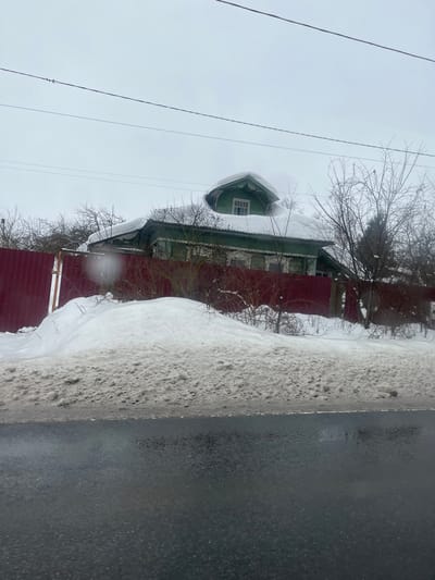 The image displays an outdoor winter scene in Podvyaznovo, Russia, under an overcast, light gray sky. A traditional green wooden house with white decorative trim around its windows and eaves stands prominently. Its roof is heavily covered in snow, from which a distinct, rounded green dormer window with white trim protrudes. A plume of white vapor or smoke is visible rising from behind the dormer. The house is enclosed by a solid red metal panel fence. In the foreground, a dark, wet asphalt road runs horizontally, bordered by a large, irregular snowdrift. The snow closest to the road is discolored with dirt and grit, while the snow closer to the fence appears cleaner. Bare deciduous trees, some with snow on their branches, are scattered around the house and behind the fence. Multiple overhead utility lines traverse the upper portion of the frame. No individuals or active subjects are present in the scene.