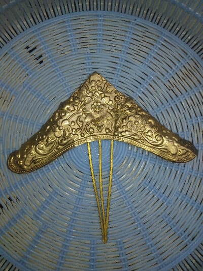 A gold-toned metallic hair ornament is depicted centrally, resting on a light blue and white radial woven surface. The ornament features a broad, curved, triangular-shaped top section, intricately embossed with floral and scrollwork designs, including a prominent central bloom. Three thin, straight prongs extend downwards from the underside of the ornate top. The background exhibits a repeating pattern of interwoven light blue and white synthetic strands, characteristic of a plastic basket or mat. No individuals or dynamic actions are visible. The scene is situated in Ottignies-Louvain-la-Neuve, Belgium.