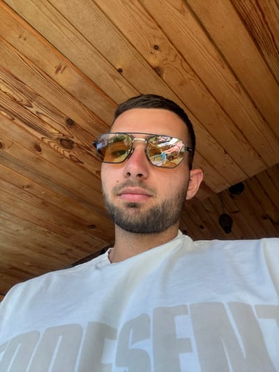 A close-up, low-angle, outdoor selfie shows a young adult male with short dark hair, stubble, and sunglasses with amber lenses. He is wearing a white t-shirt with a large, faded graphic print. The background is a wooden ceiling with visible planks and knot details. The sunglasses reflect a scene of two individuals lounging in chairs near a body of water. The image was taken in Chavei, Bulgaria, specifically on a covered patio or veranda.