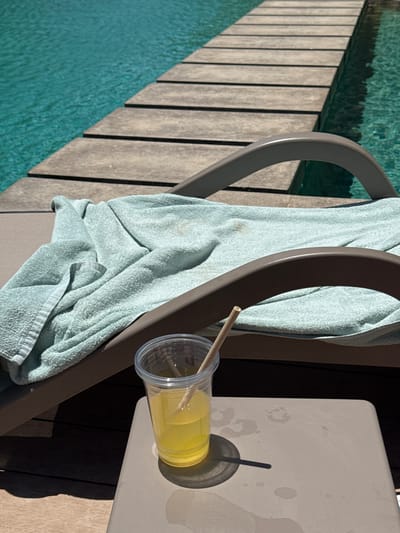 The image depicts a scene by a pool in El Gouna, Egypt. An empty plastic cup with a straw sits on a small beige table next to a lounge chair. A light blue towel is draped over the chair. The pool's edge is visible, with turquoise water adjacent to a wooden deck. The scene is brightly lit, suggesting a sunny day. No people are visible in the frame.