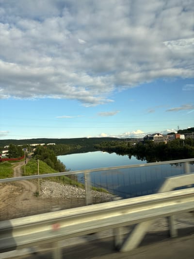This is a wide, horizontal, eye-level outdoor shot taken from a moving vehicle, likely a car or bus, on a bright, overcast day. The foreground is dominated by a metal guardrail, reflecting the environment and blurred into motion. Beyond the guardrail, a calm, reflective river flows horizontally across the frame. The river acts as a mirror, reflecting the sky, the clouds, and the trees along its banks.
On the left bank, a dirt road winds uphill through grassy slopes. A few buildings with red roofs and some smaller structures are visible near the road. Further along the left bank, a line of multi-story apartment buildings stretches along the tree line, characteristic of a residential area.
The right bank of the river is densely forested with trees that are dark green, indicating they are likely coniferous. Further inland, in the distance, there are some industrial-looking buildings with gray roofs, and tall transmission towers are visible. The sky is a vibrant blue with scattered, fluffy white clouds, some of which are quite large. Power lines stretch across the sky in several places.
The location is identified as Zverosovkhoz, Russia. The scene depicts a semi-urban environment adjacent to a natural waterway, with elements of both residential and industrial development.