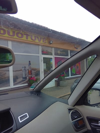The image shows a store with a sign that reads "DUOTUVE". The store appears to be a convenience store or a small market. Through the window, we can see advertisements for energy drinks and a large picture of a bottle and can of "EKSTRA" beer. There are also some potted plants outside the store. The scene is viewed from inside a car, with the dashboard and a side mirror visible. The location is Micaičiai, Lithuania.
