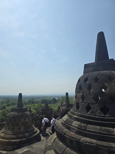 This photograph captures an aerial view of the Borobudur Temple, a Mahayana Buddhist temple located in Desa Borobudur, Indonesia. The temple's gray stone structure, characterized by its intricate diamond-shaped patterns and towering spire, dominates the foreground. Two individuals, dressed in white shirts, are positioned near the temple structure, seemingly observing the surrounding landscape. The backdrop features a vast expanse of green forest and an indistinct cityscape veiled in atmospheric haze. The sky above is a clear, light blue color.