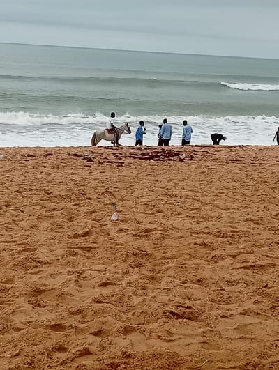 The image depicts a beach scene at Gbaj, Nigeria. A man is riding a light-colored horse near the shoreline where waves are breaking. Several other individuals are standing or bending near the horse and rider. The beach is a reddish-brown sand, and there is some scattered debris on the sand. The ocean stretches to the horizon under an overcast sky.