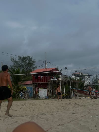A beach volleyball game is in progress in Montanita, Ecuador, at coordinates -1.825 latitude and -80.755 longitude. Three men are playing on the beach. Two men are on the court, one is preparing to spike the ball. The other is standing back, waiting for the ball to come over the net. The third man is walking towards the court, presumably to join the game. A volleyball net stretches across the court with a wooden lifeguard stand on stilts behind it, painted red and white, with a thatched roof and some items hanging on a line on the upper deck. The beach is sandy with a few trees and buildings in the background. The sky is overcast with a few scattered clouds. There is a small boat on the shore with the words "Dad y Guard" visible. The image is taken from ground level, looking up at the volleyball net and the lifeguard stand.
