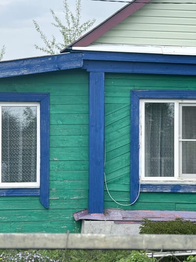 A section of a green-painted wooden building with blue trim is visible. Two windows with white frames and patterned privacy film are featured. The siding runs vertically. A white cable is attached to the building's exterior. A partial reflection of a white building and tree branches is visible in a mirror-like surface attached to the building. A concrete post and some vegetation are present on the right side of the frame. The photo appears to be taken in Ленино, Russia.