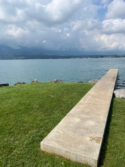 The image presents a scene along the shore of Lake Thun, Switzerland, under a partially overcast sky. In the foreground, a vibrant green, manicured lawn occupies the left and central lower portions. Extending from the lower right of the frame, a weathered, rectangular concrete slab or dock, approximately 0.5 meters wide, projects into the lake. This concrete surface displays discoloration and two faint circular depressions. Scattered grey rocks are visible along the immediate edge where the grass meets the water.

The tranquil water of Lake Thun, characterized by a clear teal-blue hue, fills the midground. Across the lake, the distant shoreline reveals a settlement of structures interspersed with trees, situated at the base of a mountain range. The mountains on the left side of the frame are higher and partially obscured by dark, heavy cloud cover. The sky above is dominated by large, white-grey cumulus clouds, with patches of clear blue visible primarily in the upper right quadrant. No individuals, animals, or active subjects are present in the scene.