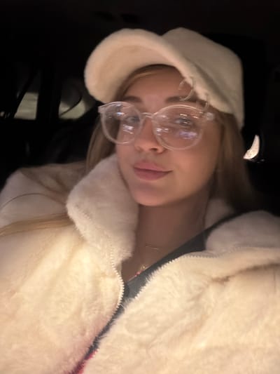 A young adult female is depicted in a close-up, chest-up shot, positioned inside what appears to be a vehicle during nighttime in the city of Азьмушкино, Russia. She has blonde hair and is wearing a cream-colored faux fur baseball cap. She wears clear, round-framed glasses, which show reflections of ambient light sources. Her lips are light-colored. She is dressed in a cream-colored faux fur jacket with a partially visible zipper. A thin, gold-colored necklace with small, colorful charms is visible around her neck. The background is dark, consistent with an interior vehicle setting at night. The subject is looking directly towards the camera.