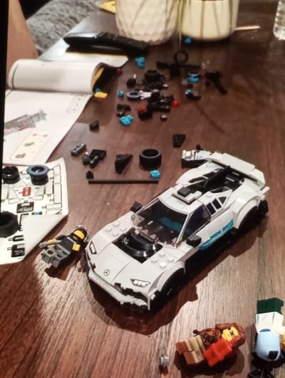 The scene is situated indoors on a wooden surface in Brzezina, Poland. The primary subject is a partially assembled Lego model of a white Mercedes-AMG sports car with black and light blue accents, positioned centrally on the wooden surface. Numerous small, unattached Lego bricks in black, grey, red, and light blue are scattered around the car. To the left of the car, an open Lego instruction manual displays assembly steps. Below the manual, a sheet of white and red Lego decals, partially used, is visible.

Three Lego minifigures are present: one grey minifigure with a yellow helmet lies prone near the front left wheel of the car. A second minifigure with a brown body, red torso, and yellow head is seated on a small brown Lego structure resembling a chair, located to the right of the car. A third minifigure, partially visible on the far right, stands upright with a light blue torso, white legs, and a green cap. In the blurred background, a dark remote control, a white textured ceramic pot, and a grey patterned fabric (likely clothing) are discernible. No direct actions or interactions between people or minifigures are depicted within the frame; the scene indicates an ongoing Lego building activity.