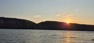 The image captures a serene sunset over the Nistru River in Molovata Nouă, Moldova. The sun, a bright orb of orange, is partially obscured by the crest of a dark, tree-covered hill, casting a golden glow and radiant beams across the water. The river's surface is rippled, reflecting the warm hues of the sky and the silhouette of the hills. The sky itself transitions from a pale blue at the top to a soft yellow and orange near the horizon, with a few wispy clouds scattered across it. The hills form a prominent silhouette in the middle ground, their slopes covered in vegetation that appears dark against the lighter sky. The overall scene is tranquil and natural, evoking the beauty of a twilight landscape.