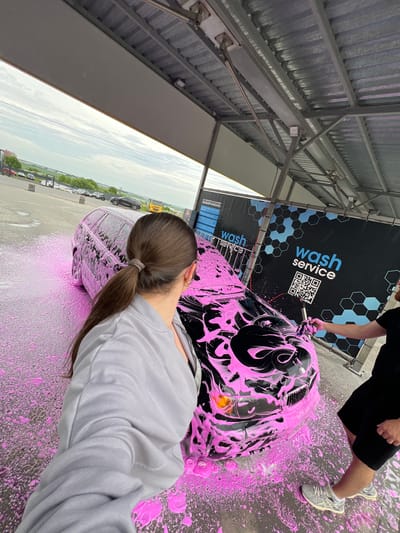 The image depicts a self-service car wash bay in Velyki Hai, Ukraine. A black BMW 3 Series station wagon is positioned centrally, heavily coated in vibrant pink cleaning foam. In the left foreground, a female with dark brown hair tied in a ponytail with a light grey band is seen from behind, wearing a light grey zippered hoodie over a lighter-toned garment and a gold hoop earring in her right ear. She holds a smartphone, indicating she is capturing the scene. To the right, a male is visible from the mid-torso downwards. He wears a black t-shirt, black shorts, white socks, and grey and white ASICS athletic shoes. He is actively applying or spreading the pink foam on the front of the BMW with a blue sponge/brush. The concrete ground surrounding the car is wet and covered with dripping pink foam. The car wash structure features a corrugated metal roof supported by grey metal beams. On the back wall, two black banners display 'wash service' branding, light blue hexagonal patterns, and a QR code. The banner on the left partially shows Ukrainian text: "НА ТЕРИТОРІЇ АВТОМИЙНОЇ ЗАБОРОНЕНО:". Beyond the car wash bay, an open outdoor area is visible with a paved surface, parked vehicles (including a yellow truck), green vegetation, and an overcast sky.