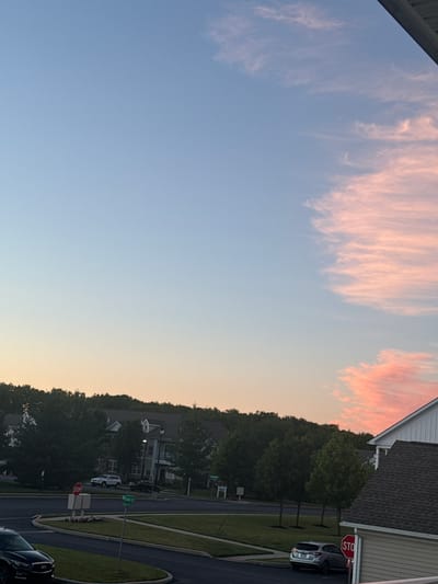 The image shows a suburban street scene in Allen Township, United States of America, during a sunset. The sky is a gradient of blue and pink, with wispy clouds illuminated by the setting sun. In the foreground, a dark sedan is parked. The middle ground features a street with a stop sign, a street sign reading "Wilson Ct / Rosa Ct," and a white SUV. Beyond the street, a line of trees forms a backdrop to residential buildings with pitched roofs. A portion of a house with a dark shingled roof is visible in the right foreground. The overall impression is of a peaceful evening in a residential area.