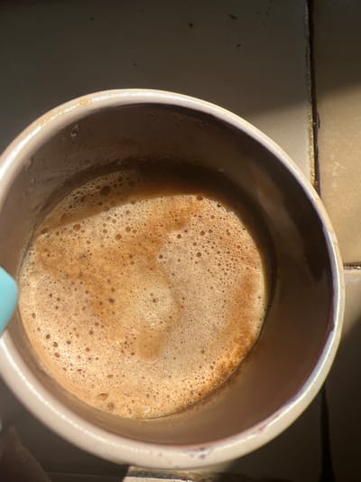 A close-up, overhead shot captures the surface of a frothy beverage, likely coffee, within a light-colored ceramic mug. The liquid has a creamy, light brown color with a thick layer of small, uniform bubbles. Sunlight streams in from the right, creating a diagonal highlight across the foam and casting shadows on the mug's rim and the tiled surface beyond. The background appears to be a countertop with beige tiles and dark grout lines. There is a hint of a light blue object, possibly a spoon or part of another utensil, entering the frame from the left. The overall impression is one of a casual, sunlit moment. The location is described as being in the city of Karabunar, Bulgaria.