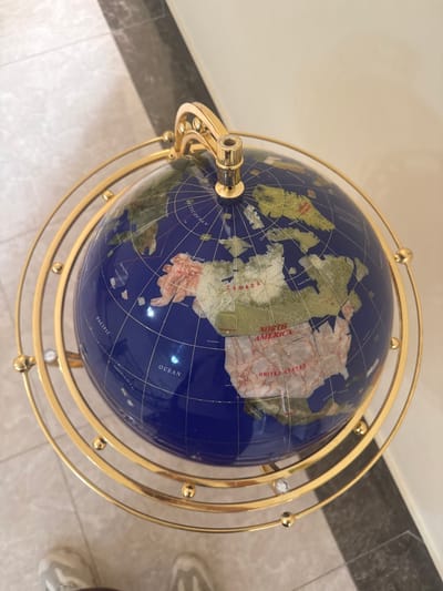 The scene depicts an ornate decorative globe situated indoors on a polished floor in Aragats, Armenia. The globe features deep blue oceans, likely crafted from lapis lazuli or a similar stone, with continents formed from various cut and polished natural stones. Notable landmasses include North America, with Canada rendered in a light green stone and the United States appearing in a reddish-pink stone. Text labels such as "NORTH AMERICA," "UNITED STATES," "CANADA," "ARCTIC OCEAN," "PACIFIC OCEAN," and "OCEAN" are visible. The globe is mounted within a polished golden or brass-toned metal armillary sphere-style stand, comprising multiple concentric rings. This stand is embellished with small golden spheres and at least two clear, faceted stones. A golden finial serves as the globe's top axis point. The floor beneath the globe consists of light-colored, possibly beige or grey, tiles. A darker tile border runs along the right side, adjacent to a plain, light-colored wall or vertical surface. In the lower left corner, the tips of two human feet wearing light-colored athletic shoes are visible.