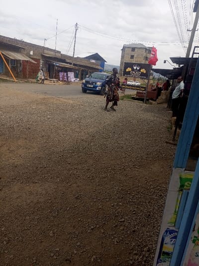 The image depicts an unpaved street scene in Kiamaina ward, Kenya, under an overcast sky. The ground in the foreground and middle ground consists of dark earth mixed with light-colored gravel. A male individual, dressed in dark clothing, walks from right to left across this unpaved area, carrying several bundles of fabric or clothing draped over his arm and shoulder.

To the left, a blue Toyota Hilux pickup truck with license plate KBY 591T is present on the unpaved road, positioned facing slightly towards the right foreground. Behind the truck, a series of single-story commercial structures with corrugated metal roofs line the street; one displays purple and white merchandise (possibly chairs or furniture) outside its entrance. Further left, another person wrapped in light-colored fabric stands near one of these buildings.

In the background, a multi-story stone building with multiple windows is visible. A red flag, featuring a white emblem resembling an Arsenal FC cannon, flies from an upper portion of this building. Below it, a large black sign displays the word "ENJOY" in gold lettering, accompanied by an image of two clinking wine glasses. To the far right, partially visible blue wooden structures, likely part of a market stall or shop, frame the foreground, with merchandise bags visible at the bottom right. Utility poles and overhead power lines traverse the scene against the grey sky. Distant hills or foliage are faintly visible on the horizon.