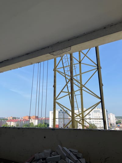 The image depicts a view through an unfinished concrete structure, likely a building under construction or renovation, in Аснежыцкі сельскі Савет, Belarus. In the foreground, a rough concrete ledge occupies the bottom portion of the frame, with a pile of construction debris, including broken concrete blocks, visible on it. Above this, a bright blue sky serves as the backdrop. Dominating the middle ground is a large, yellow metal structure, appearing to be part of a crane or scaffolding, with visible ladder rungs. Several thin, dark vertical wires or cables hang from the concrete overhang in the upper portion of the image, obscuring parts of the sky and the yellow structure. Beyond the yellow structure, a cityscape unfolds, characterized by rows of apartment buildings with red and gray roofs, interspersed with trees. The buildings vary in height and architectural style, suggesting a mix of older and newer constructions. The overall scene is one of ongoing construction within a partially built environment, offering a distant view of the surrounding urban landscape.