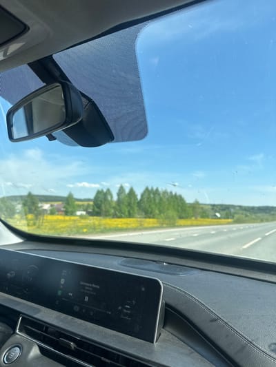 The image is taken from inside a car, looking out through the windshield. The dashboard and rearview mirror are visible. Outside, the car appears to be driving on a highway or road. There is vegetation on the side of the road, including trees and yellow wildflowers. The sky is blue with some clouds. The car's infotainment system is visible, displaying a music track titled "Llorona Remix" by Santton. The image does not include enough information to determine if the location is in the city of Postol, Russia.