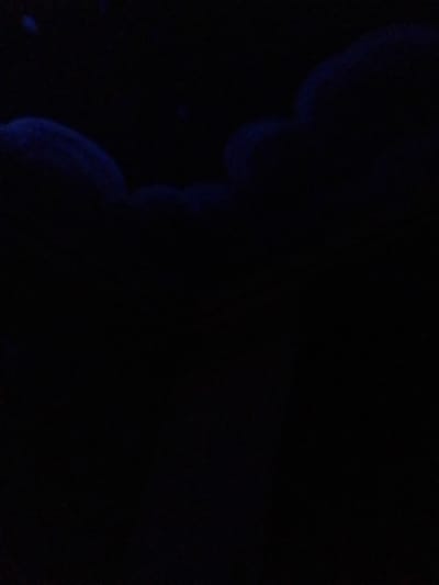 The image displays an extremely dark scene, predominantly black, with faint, indistinct blue illumination visible across the upper portion. Curved, formless blue shapes or light sources are discernible in the top third of the frame, providing minimal ambient light against the deep shadows. No discernible people, objects, structures, or specific environmental features are present. The specific location within the city of Long Beach, United States, cannot be determined from this image.