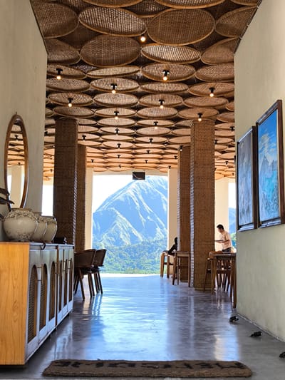Captured from a low-angle perspective, the photograph showcases the interior of a building in Sembalun Bumbung, Indonesia. The flooring is a polished gray, reflecting ambient light. A brown mat sits at the entryway. To the left, a light-brown wooden cabinet holds three large ceramic vases. An oval mirror hangs above the cabinet. Wicker chairs are present. The ceiling is adorned with woven round baskets, each housing a small light fixture. Tall columns with a woven design support the ceiling. At the end of the corridor, a mountain is visible through a large opening. Two people are seated at tables on either side of the corridor. A man in a tan shirt is standing, holding a tray. Two framed pictures are hanging on the wall to the right.