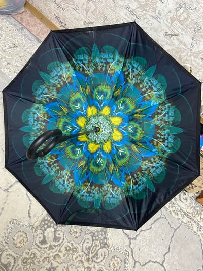 The image shows an opened umbrella with a vibrant, circular feather pattern. The pattern features shades of blue, green, and yellow, resembling peacock feathers. The umbrella is black around the edges and has a black curved handle. It is lying open on a floor covered with a patterned carpet. In the background, there is a wall with a pattern resembling vintage wallpaper featuring French text like "Bonjour" and "Paris". The setting appears to be indoors. There are no people or animals visible in the image. The location is specified as Бескөл ауылдық округі, Kazakhstan, though this information is not visually discernible from the image itself.