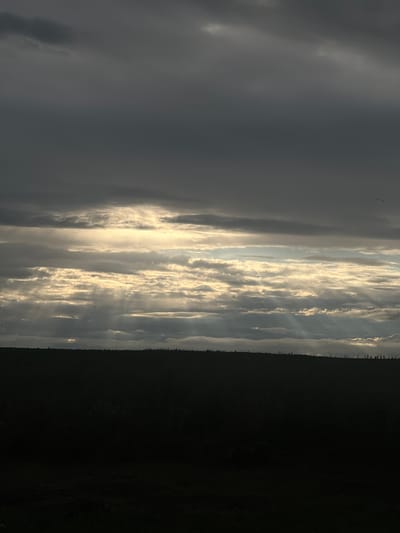 A landscape image depicts a dark, silhouetted field under a dramatic, overcast sky. The clouds are a mix of dark grays and lighter grays, with some areas allowing shafts of golden sunlight to break through, creating a layered effect in the sky. The horizon line is a stark contrast between the dark foreground and the illuminated sky, with the silhouette of vegetation along the top edge of the field. The overall mood is somber and atmospheric, with the play of light and shadow creating a sense of depth and drama. The image was taken in Бучацінскі сельскі Савет, Belarus.