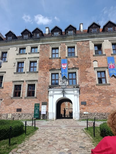 The image shows the Grand Dukes Castle in Uniejów, Poland. The castle is a red brick building with a large arched entrance in the center. Two blue banners with coats of arms are hanging from the facade. The castle is surrounded by a courtyard paved with cobblestones. There is a person standing in the arched entrance, and another person in the foreground on the right. The sky is blue with some clouds.