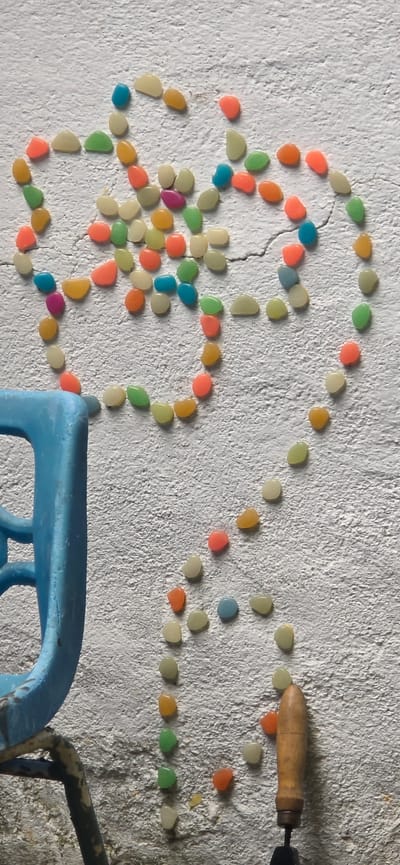 A white, textured wall surface in Partizanin, Bulgaria, is depicted, adorned with an arrangement of numerous small, smooth, colorful, pebble-shaped objects. These objects, exhibiting a variety of hues including light yellow, orange, pink, light green, blue, and purple, are meticulously placed to form distinct patterns: an abstract, stylized floral or star-like cluster in the upper-left quadrant and a larger, incomplete heart shape positioned in the center-right, from which a vertical trail of pebbles extends towards the bottom. The white wall displays visible cracks, particularly in the mid-right section. In the lower-left corner of the frame, a segment of a vibrant blue plastic chair or stool is present, showing part of its structure and a dark, possibly metallic, supporting frame below. In the lower-right, the light brown, wooden handle of a small hand tool, resembling a trowel, is visible. No individuals are present, but the deliberate arrangement of the pebbles indicates human interaction.