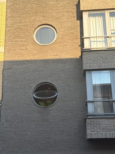 The image shows a close-up, exterior view of a modern brick building with two distinct styles of brickwork. The upper portion of the building features light brown, textured brick, while the lower section is constructed with darker gray, rectangular bricks. Two circular windows are visible: one near the top of the light brown section, which is a closed, circular pane of glass, and another in the dark gray section, which is a circular opening with a hinged section of glass propped open, revealing greenery inside. To the right of the circular windows, a modern section of the building with large, rectangular windows and a balcony is visible. The overall impression is one of contemporary architecture with unique window designs, located in Strombeek-Bever, Belgium. There are no people or animals present in the image. The lighting suggests it is daytime, with sunlight casting shadows on the building's facade.