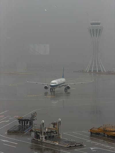 An airplane sits on a wet tarmac at an airport in Guangyang District, China, under a heavy fog. The airplane, with blue and white livery, is positioned on a taxiway, facing forward. In the background, a distinctive airport control tower with a white, ribbed structure rises into the hazy sky. Several airport service vehicles, including baggage carts and loaders, are present on the tarmac in the foreground. The ground is slick with water, indicating recent or ongoing rain. The overall scene conveys a sense of a quiet, possibly delayed, operational day due to the weather conditions.