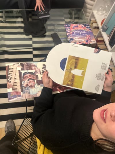 A person holds a white vinyl record with a blue center and a white jacket featuring a yellow and white image. Below, two other album covers are visible: one a collage of faces and a keyboard, the other a colorful abstract design. The scene takes place on a glass table with a black and white striped rug underneath. A bookshelf filled with books and glass objects is partially visible in the background. The person wears a black sweater and stands near a black metal chair.