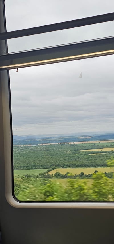 The image shows a view from a train window overlooking a landscape. The foreground features blurred green foliage, indicating motion. Beyond this, there is a vista of rolling hills, dense green forests, and lighter green fields, possibly farmland. In the distance, the landscape fades into hazy blue hills under a cloudy, overcast sky. The sky contains a small, triangular white object, possibly a distant kite or an aircraft. The window frame, made of dark grey material, is visible along the edges of the image, with a dark horizontal bar and a light strip above the window. There is no indication of the location being Kozar Belene, Bulgaria.