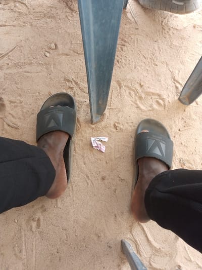 The scene, located in Orudu-Olokan, Nigeria, presents a top-down view of an individual's lower legs and feet on a light brown, sandy ground. The person wears black trousers and dark grey slide sandals, each featuring a raised geometric logo and the inscription "MADE IN TURKEY" on the strap. Between the feet, a small, crumpled pink and white wrapper with partial text "industry" lies on the sand. Multiple dark grey or blue metallic chair legs are visible, partially sunk into the sandy surface, suggesting the individual is seated or standing amidst outdoor furniture. The sand exhibits minor irregularities and scattered small debris. No other individuals are present in the frame.