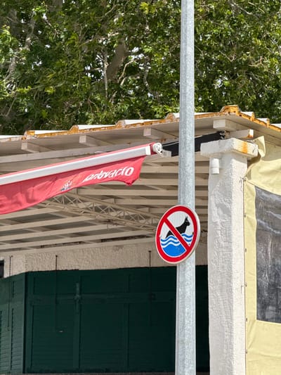 A metal pole with a red and blue "no dogs" sign is mounted on the side of a building. The sign shows a black silhouette of a dog swimming. The sign is mounted in front of a green door and under a red awning that says "Lavoracio" on a white building. There is a tree in the background. The scene is in Dugi Rat, Croatia. The location, based on coordinates (latitude: 43.446, longitude: 16.634), is near the waterfront.