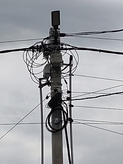 The image shows a utility pole with numerous electrical wires and a small rectangular electronic device mounted at the top. The pole is made of concrete and is situated outdoors against a cloudy sky. Several bundles of black electrical wires are wrapped around the pole and extend outwards. A small, gray box with the number "7437" printed on it is attached to the pole, along with other electrical components and mounting brackets. The overall impression is one of a densely wired urban or semi-urban environment, possibly related to telecommunications or power distribution. The location is specified as Skurygino, Russia.