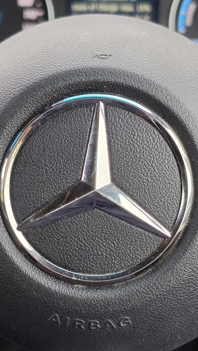 The image shows a close-up of a Mercedes-Benz steering wheel. The central feature is the Mercedes-Benz logo: a three-pointed star within a circle, rendered in polished metal. The surrounding material of the steering wheel is black and textured, presumably leather or a synthetic substitute. The word "AIRBAG" is partially visible on the right side of the image, indicating the presence of an airbag system. The image does not provide information regarding the location of the city of Bolton, United Kingdom.