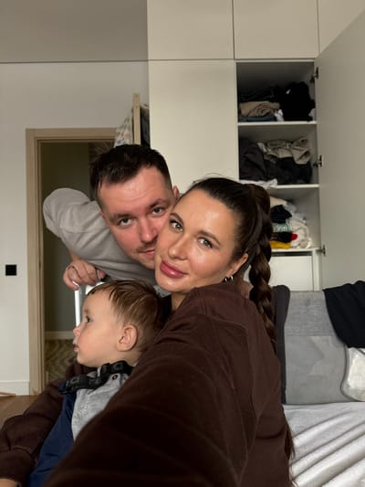 A domestic interior scene captures three individuals: an adult female, an adult male, and a young child. The scene is situated in the city of Gribovo, Russia, as per the provided context.

The adult female, with dark hair tied in a high ponytail and a visible braid, has a light complexion and light-colored eyes. She wears a brown long-sleeved top and a silver hoop earring in her visible left ear. She is positioned towards the center-right, looking directly towards the camera with a gentle smile. She appears to be holding the camera.

The adult male, with short dark hair, light complexion, and light-colored eyes, has stubble on his face. He wears a light gray long-sleeved shirt and a gold-colored band on his left ring finger. He leans in from behind the female on the left, looking directly at the camera with a slight smirk. His right hand is visible holding onto a lighter-colored object, possibly a part of a bed frame or furniture.

The young child, of toddler age, has light brown hair and is positioned in front of the female, largely obscured but facing left, away from the camera. The child wears a grey long-sleeved shirt layered over a navy blue one, with a black strap accessory around the collar area.

The background consists of light-colored walls. To the right, a white modular cabinet unit is visible, with its top-right door open, revealing folded clothes of various colors (black, beige, white, red, green). To the left, a light-colored wooden door frame and a dark door handle are present, with a glimpse of a light wooden floor through the doorway. A white textured blanket or sheet covers a surface, likely a bed or sofa, in the bottom right, next to a gray pillow. The lighting is even, suggesting an indoor ambient light source.