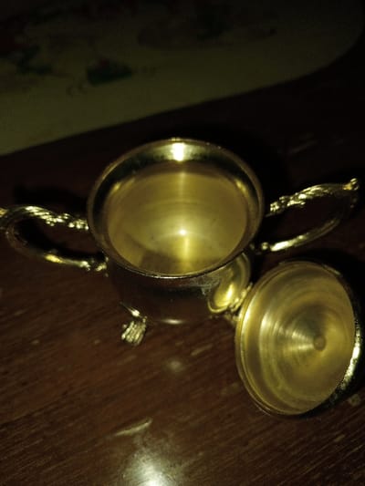 The image displays a small, ornate, golden-colored metal pot or creamer with a detached lid, positioned on a dark brown wooden surface in Longmiang, Laos. The pot features two twisted, decorative handles and rests on three small, intricately sculpted feet. Its interior is visible and empty, exhibiting a reflective golden surface. The lid, also golden-colored, lies to the right of the pot, revealing its inner surface which has concentric circular patterns. The wooden surface beneath the objects shows visible grain and reflects some light. The background is dimly lit and out of focus, showing indistinct shapes and muted colors, possibly a patterned surface. No individuals or discernible actions are present in the scene.
